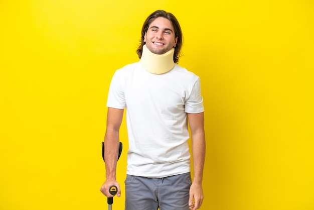 Young handsome man wearing neck brace and crutches isolated on yellow background looking to the side and smiling