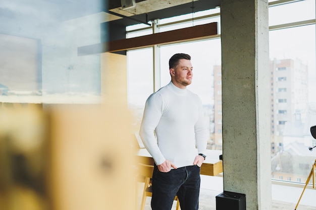 A young handsome man in a white sweater is standing in a spacious bright office The concept of a modern successful person Young smiling guy in open office space