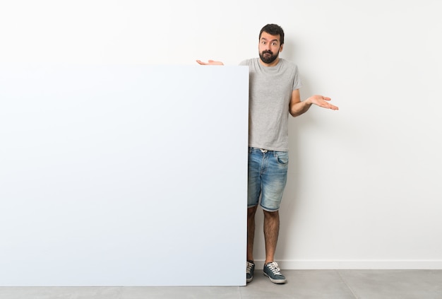 Young handsome man with beard holding a big blue empty placard having doubts while raising hands