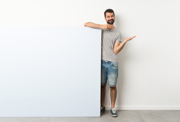 Young handsome man with beard holding a big blue empty placard holding copyspace imaginary on the palm to insert an ad