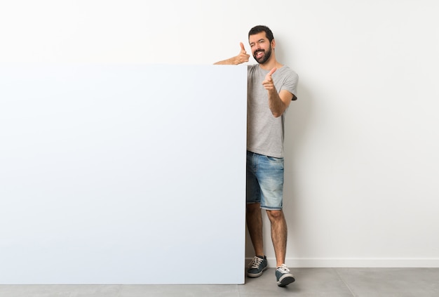 Young handsome man with beard holding a big blue empty placard pointing to the front and smiling