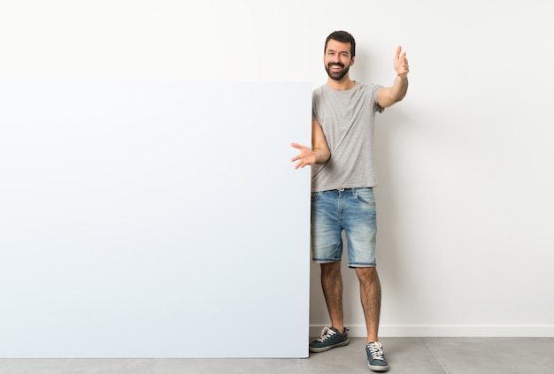 Photo young handsome man with beard holding a big blue empty placard presenting and inviting to come with hand