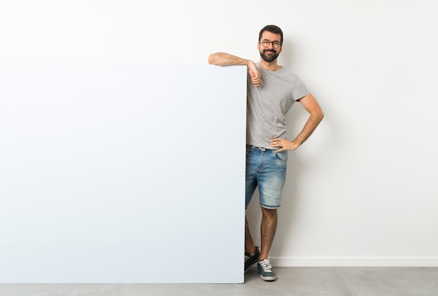 Young handsome man with beard holding a big blue empty placard with glasses and happy