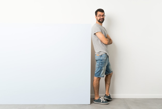 Young handsome man with beard holding a big blue empty placard with glasses and smiling