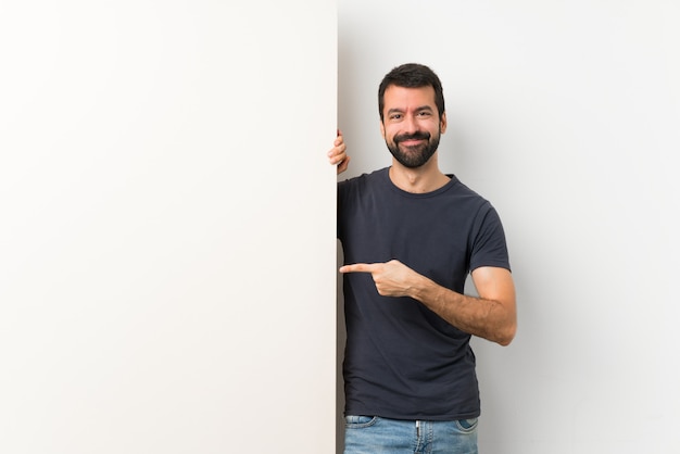 Photo young handsome man with beard holding a big empty placard pointing finger to the side