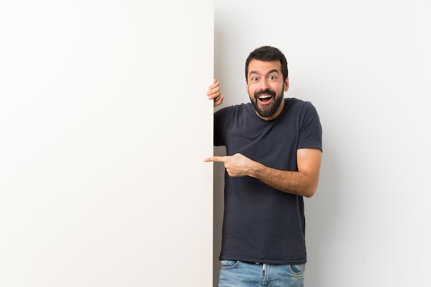 Photo young handsome man with beard holding a big empty placard surprised and pointing finger to the side