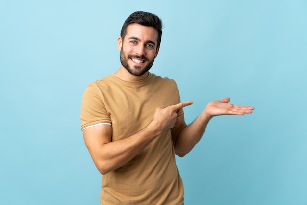 Young handsome man with beard over isolated wall holding copyspace imaginary on the palm to insert an ad