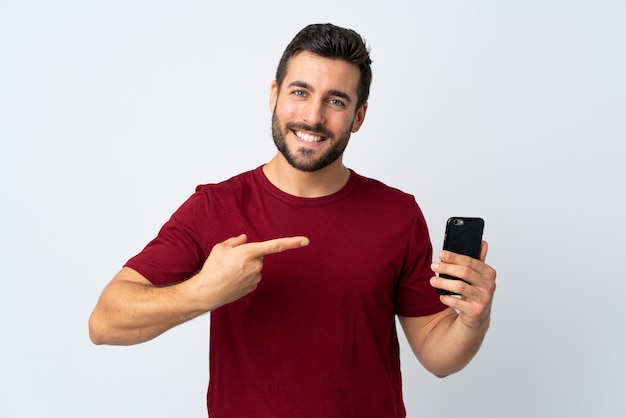 Young handsome man with beard using mobile phone isolated on white wall and pointing it