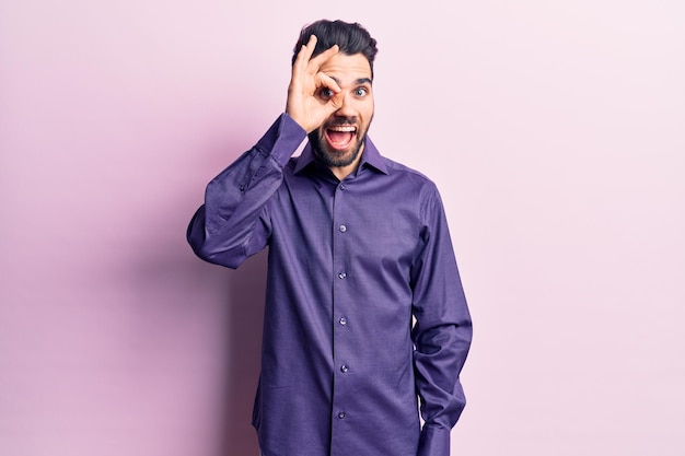 Young handsome man with beard wearing casual shirt doing ok gesture with hand smiling, eye looking through fingers with happy face.