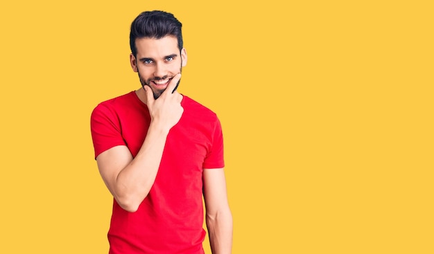 Young handsome man with beard wearing casual t-shirt looking confident at the camera smiling with crossed arms and hand raised on chin. thinking positive.