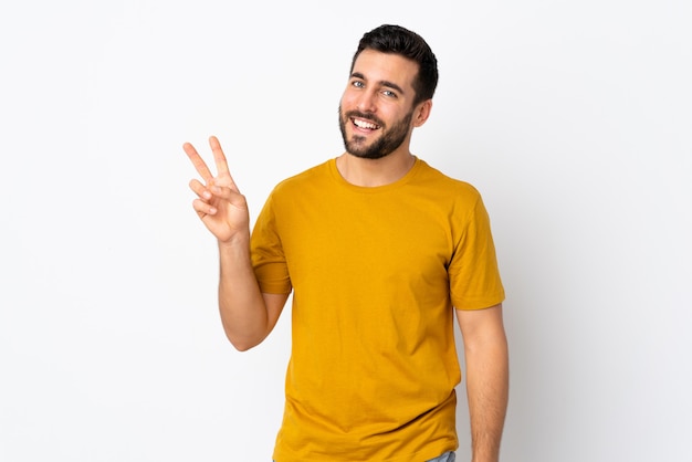 Young handsome man with beard on white wall smiling and showing victory sign