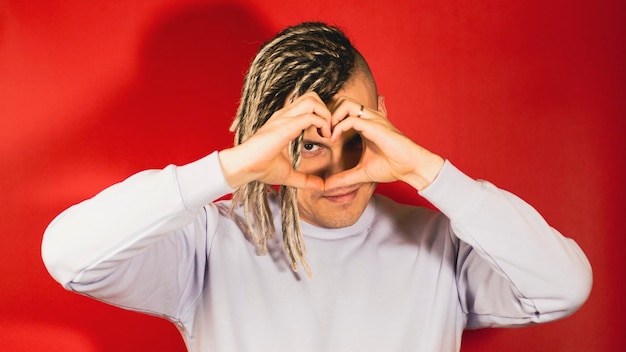 Young handsome man with blonde dreadlocks showing heart with hands Attractive smiling guy looks at camera through heart gesture with hands on red background