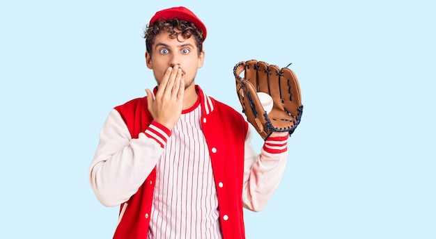 Young handsome man with curly hair wearing baseball uniform holding golve and ball covering mouth with hand shocked and afraid for mistake surprised expression