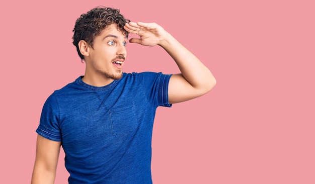 Young handsome man with curly hair wearing casual clothes very happy and smiling looking far away with hand over head. searching concept.