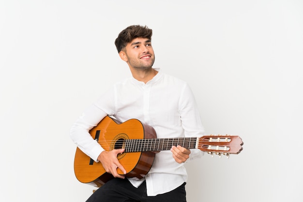 Young handsome man with guitar over isolated white wall looking up while smiling