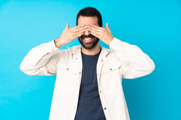 Young handsome man with white corduroy jacket over isolated blue wall covering eyes by hands