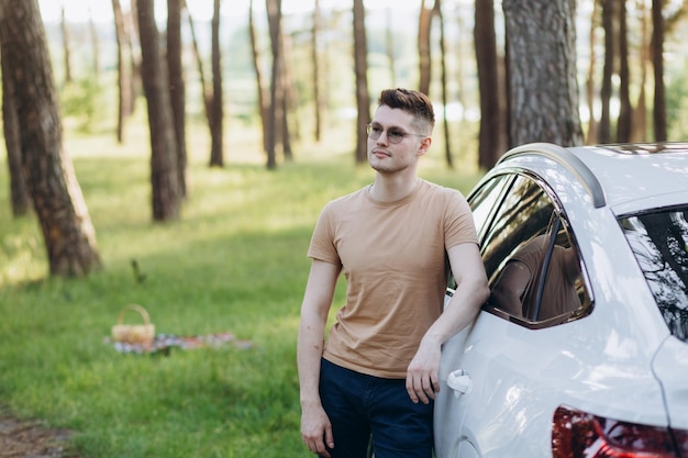 Young handsome smiling man holding charging cable at electric charging station point standing near his new car looking satisfied