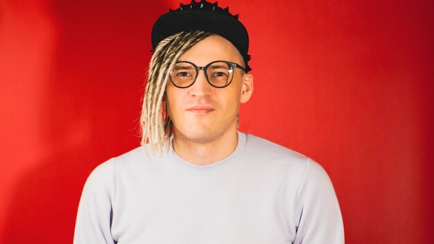 Photo young handsome stylish man in glasses in black cap on red background hipster looking at camera with blonde dreadlocks