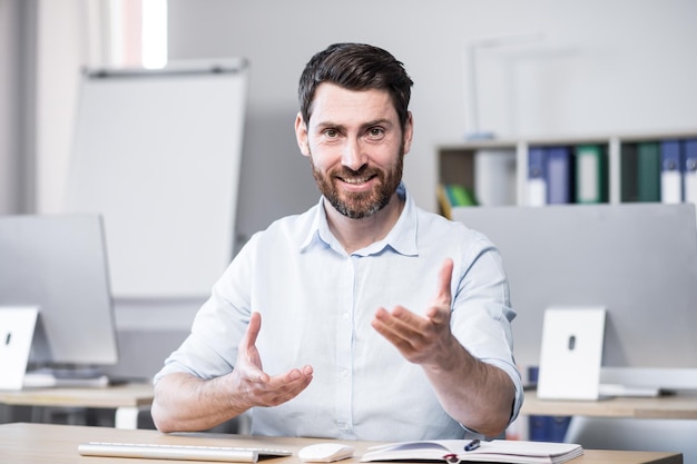 Young handsome successful business coach explains and tells on camera gestures with his hands Sitting at a computer desk in the office looking at the camera