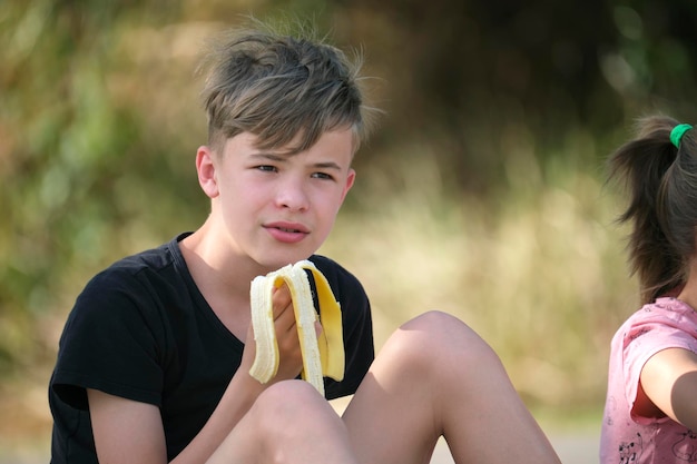 Young handsome teenager boy eating tasty ripe banana snacking outdoors on summer day