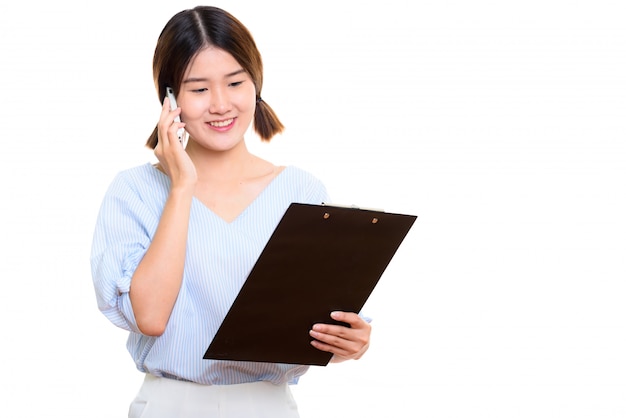 Young happy Asian woman smiling while reading on clipboard and talking on mobile phone