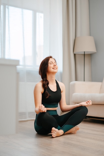 Young happy athlete in the lotus position is sitting on the floor of the house doing yoga