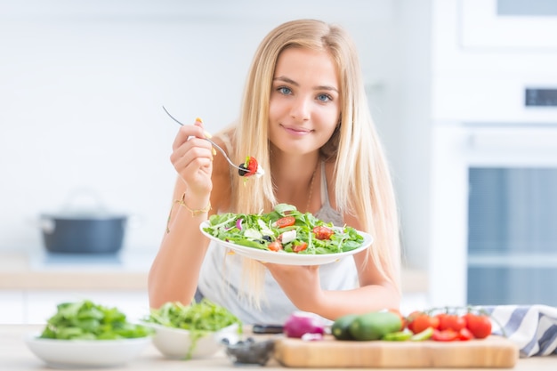 Young happy blonde girl eating healthy salad from arugula spinach tomatoes olives onion and olive oil.