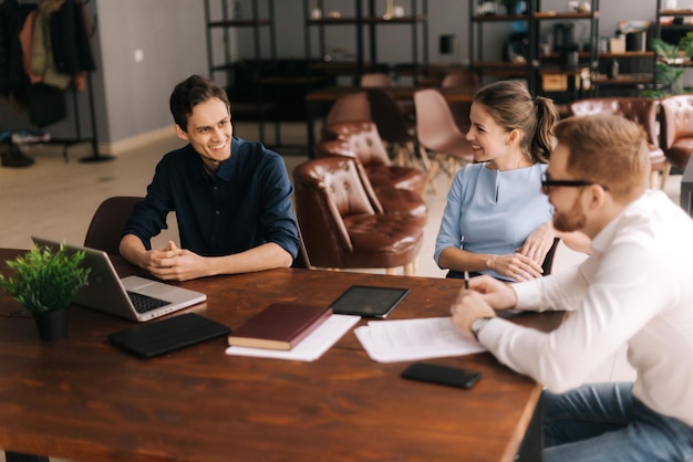 Young happy boss explaining project strategy to executive manager in modern office meeting. Colleague listen to the speaker. Young man sits at the table with his laptop.Concept of office life.