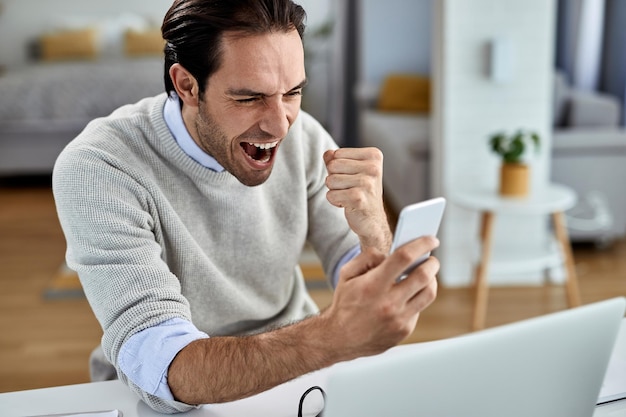 Young happy businessman using smart phone and celebrating good news while working at home