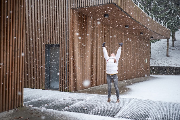 Young happy caucasian girl with pink backpack dressed in white jacket smiling and enjoying first snow with rised hands