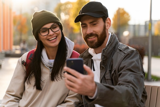 Young happy caucasian lover woman and man embracing together tenderly while sitting at the bench at the street and making selfie with each other. Love and date concept