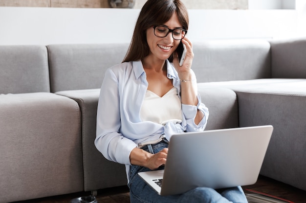 young happy cheerful business woman indoors at home on sofa in living room using laptop computer talking by mobile phone.