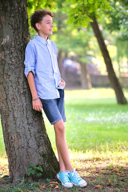 Young happy child boy relaxing in summer park leaning on tree trunk Child wellbeing concept