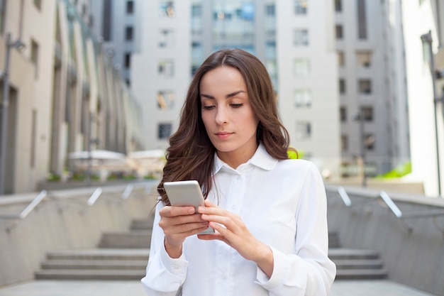 Young happy confident business woman is using her smartphone while walking outdoors in an urban city