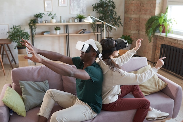 Young happy couple in glasses sitting on sofa and gesturing while playing virtual reality game at home