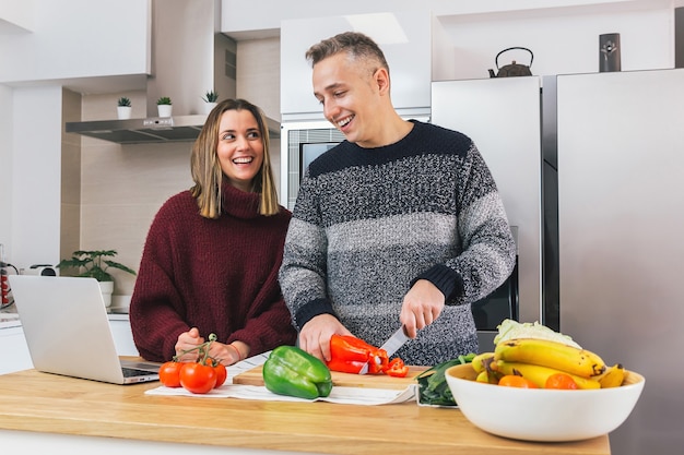 Young happy couple is laughing and preparing healthy food in their kitchen and reading recipes on the notebook