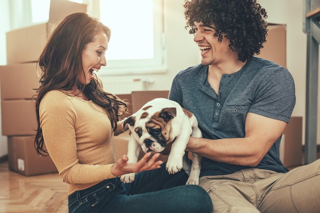 The young happy couple is moving into a new house. They are holding their little puppy after they brought boxes with things to their new home.