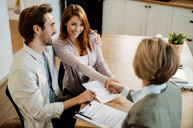 Young happy couple shaking hands with insurance agent while having a meeting at home Focus is on young woman