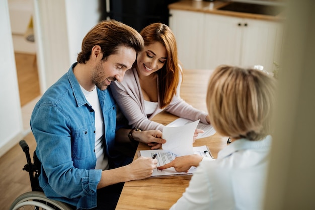 Young happy couple signing a contract with real estate agent on a meeting at home. Focus is on man in wheelchair signing a document.