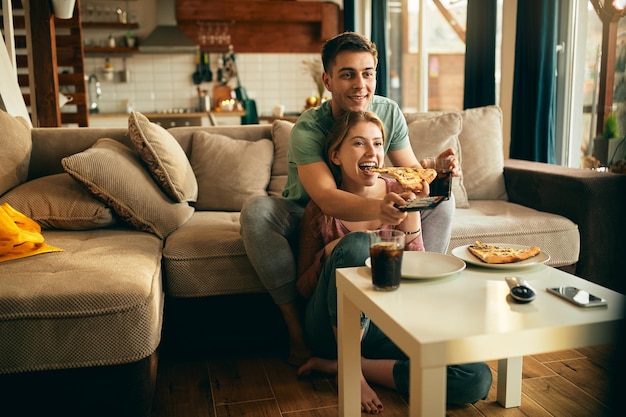 Young happy couple watching TV while eating pizza in the living room