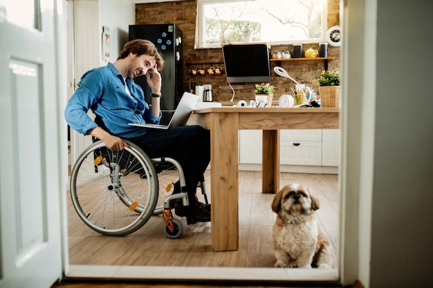 Young happy entrepreneur in wheelchair surfing the net on laptop while being with his dog at home