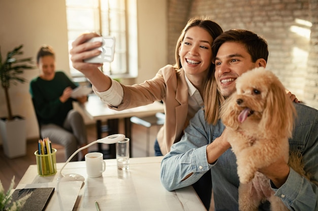 Young happy entrepreneurs having fun while taking selfie with a poodle in the office