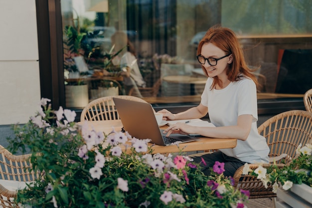 Young happy european woman sitting at cafe table outdoors with laptop and cup of coffee, smiling redhead female in eyeglasses enjoying remote work at coffee shop or studying online