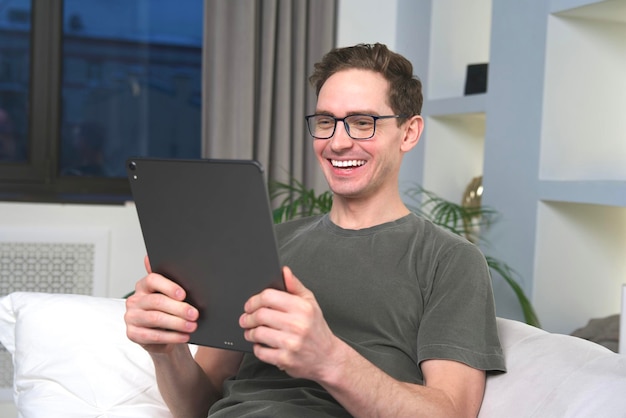 Young happy excited man in glasses is smiling celebrating success victory win looking at screen of tablet computer at home in living room sitting on couch or sofa Good news happiness