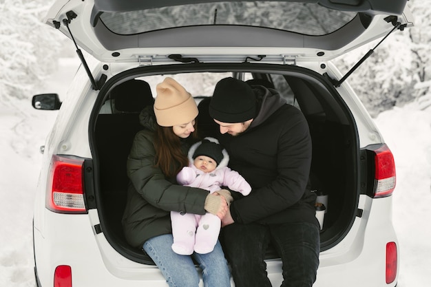 Young happy family a man a woman and a baby are sitting in the trunk of a car in a winter