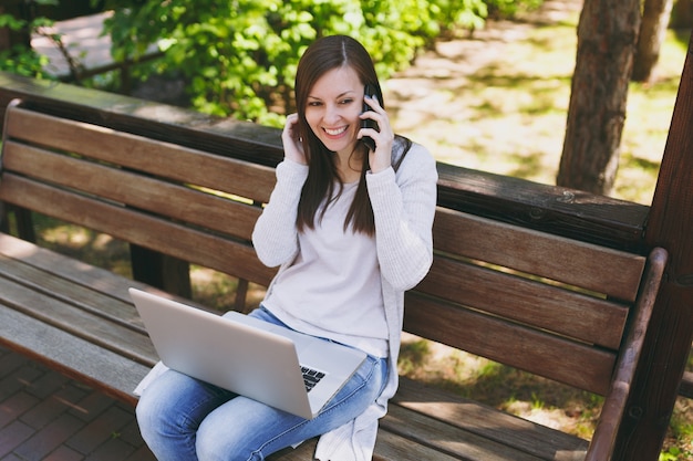 Young happy female in light casual clothes talking on mobile phone. Woman sitting on bench working on modern laptop pc computer in street outdoors on nature. Mobile Office. Freelance business concept.