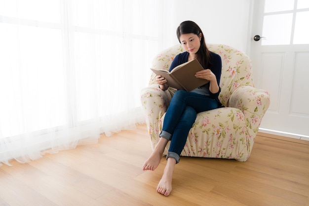 young happy girl studying knowledge book at free time sitting on living room comfortable sofa in front of window relaxing.