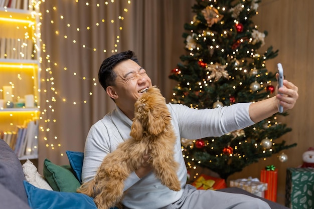 Young happy handsome asian man holding phone and taking selfie with his dog sitting at home on