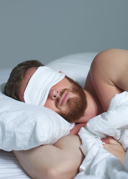 Young happy handsome sleepy man in the bed in bedroom at home in the morning lying under white blanket enjoy resting sleep well in night mask on face eyes Vertical photo