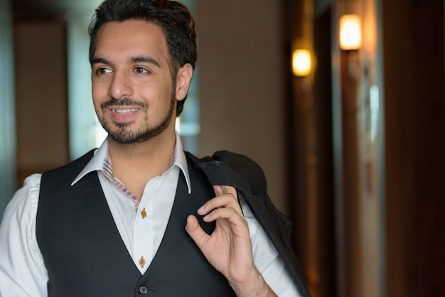 Young happy Indian man smiling while holding jacket and looking away in the corridors of hotel